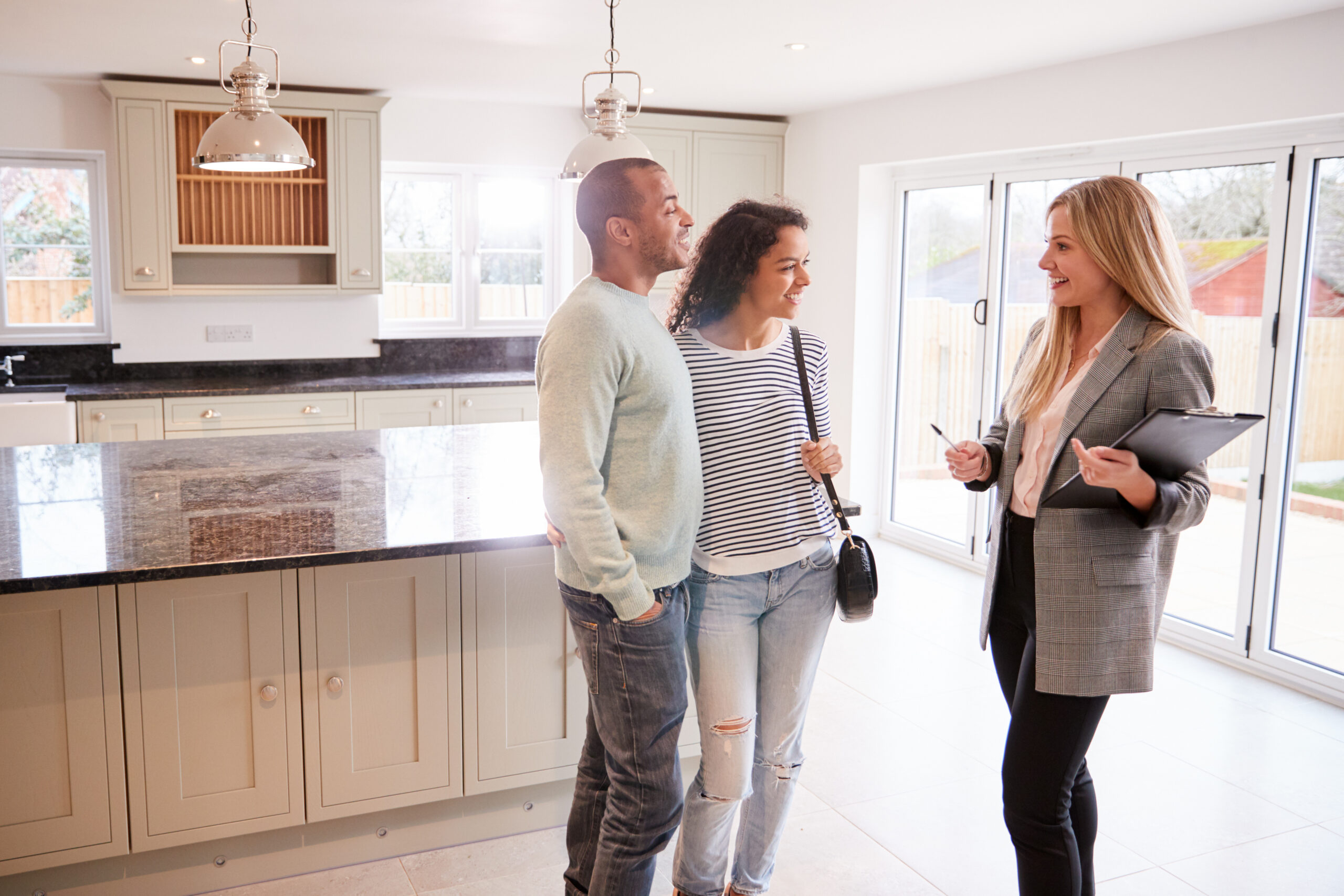 A young couple (man and woman) are speaking to a woman REALTOR in the kitchen area of a home. There is a kitchen island and sliding glass doors providing bright lighting in the room.